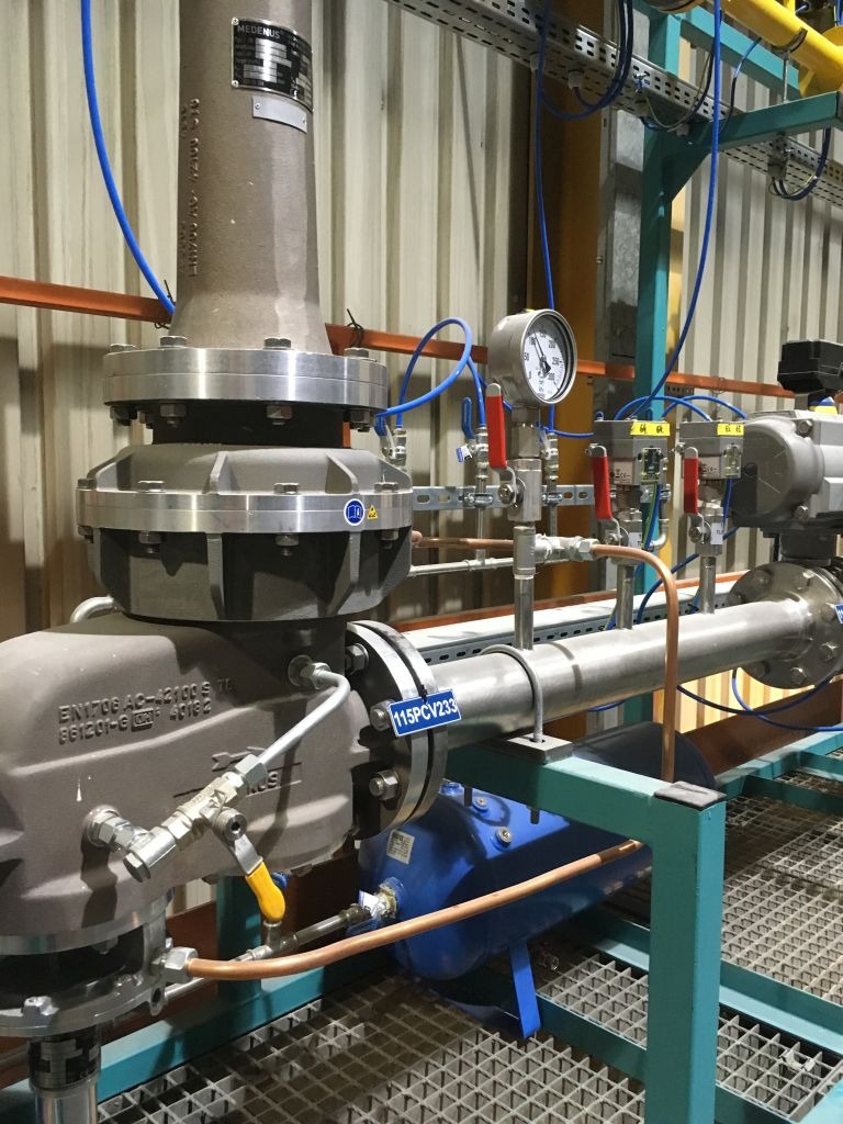 Kompleksowa instalacja przemysłowa z grubymi rurami, zaworami z żółtymi uchwytami, manometrem, niebieskimi przewodami i niebieskim zbiornikiem, zamontowana na metalowej konstrukcji kratowej, w tle...