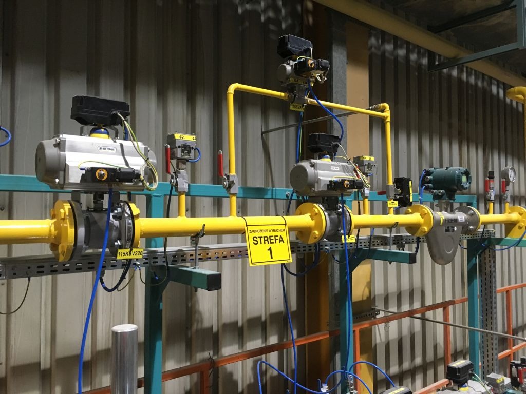 Żółte rury gazowe z zaworami i czujnikami w przemysłowej instalacji, tabliczka ostrzegawcza 'Zagrożenie wybuchem STREFA 1'.