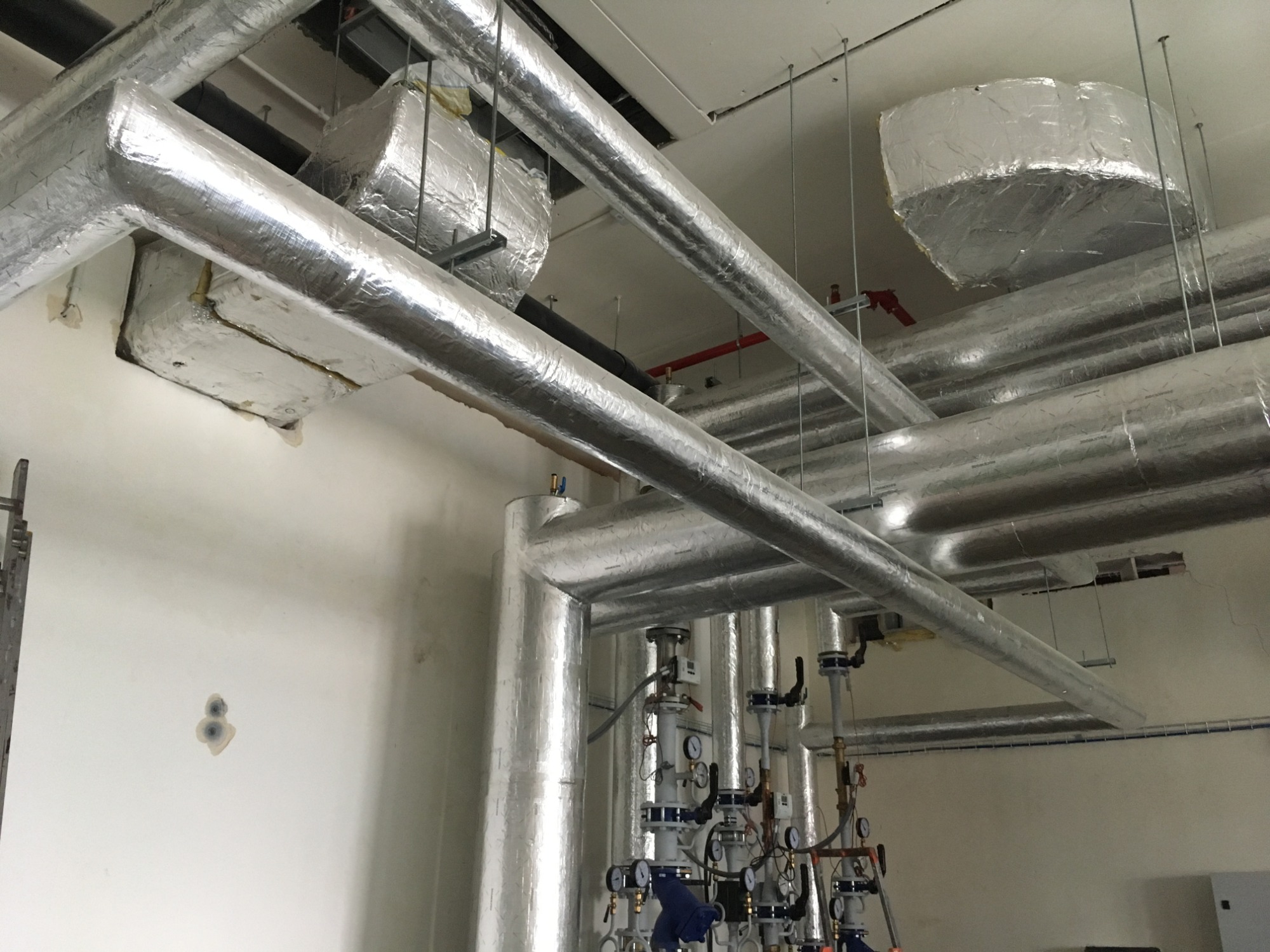 Izolowane rury i elementy instalacji przemysłowej pod sufitem, z widocznymi zaworami, manometrami i elementami mocującymi, odzwierciedlające światło w folii izolacyjnej.