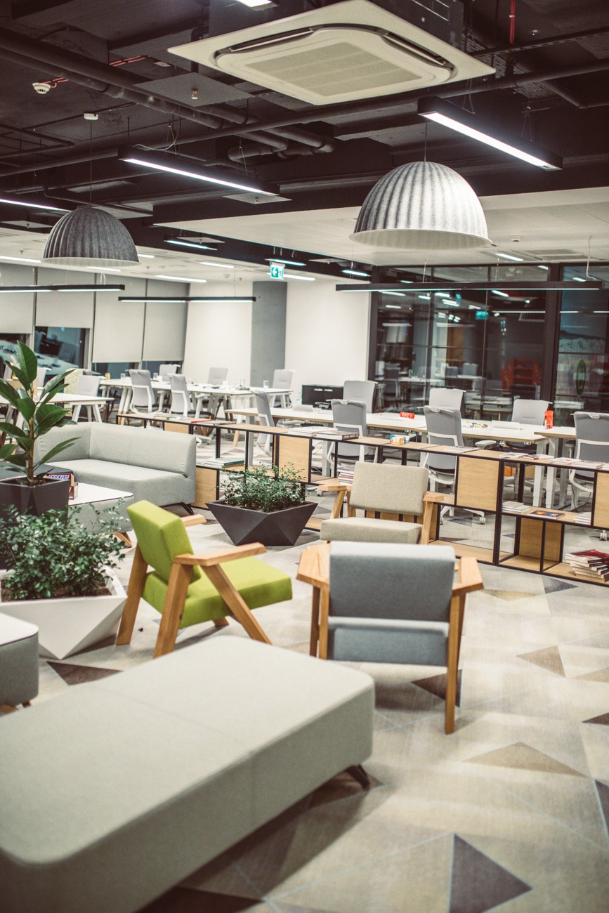 Wnętrze biura z sufitowymi lampami w kształcie kopuł, rzędami biurek z szarymi krzesłami, roślinami w geometrycznych donicach oraz meblami wypoczynkowymi w odcieniach szarości i zieleni.