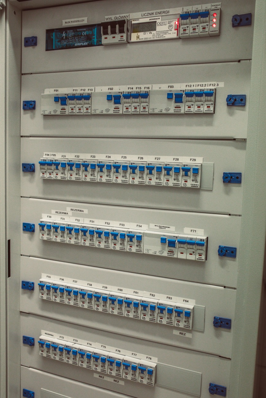 Szczegółowe ujęcie rozdzielnicy elektrycznej z modułowymi wyłącznikami, licznikiem energii i blokiem rozdzielczym, widoczne oznaczenia F01-F78 oraz napisy 'REZERWA', 'WYŁ GŁÓWNY', 'LICZNIK ENERGII'...