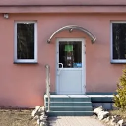 Fasada biurowca w odcieniach różu z białymi oknami i drzwiami wejściowymi, przed wejściem mała choinka, tabliczka informacyjna po prawej stronie od wejścia.