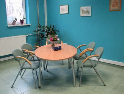 Jasne biuro z okrągłym stołem, krzesłami z drewnianymi podłokietnikami, kwiatami w wazonie i dekoracjami na ścianach, sugerujące przestrzeń do spotkań z klientami.