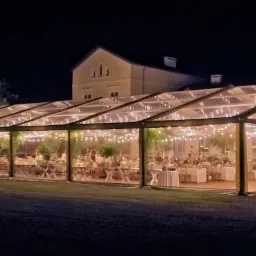 Przezroczysty namiot eventowy z oświetleniem girlandami, ustawiony na trawniku przed budynkiem, przygotowany do organizacji przyjęcia.