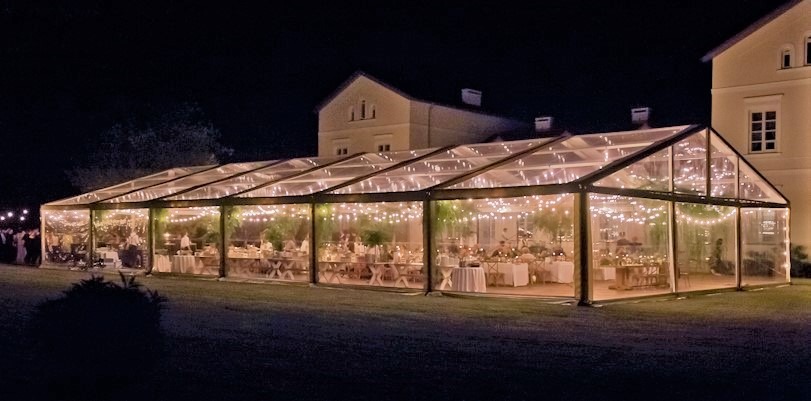 Przezroczysty namiot eventowy z oświetleniem girlandami, ustawiony na trawniku przed budynkiem, przygotowany do organizacji przyjęcia.