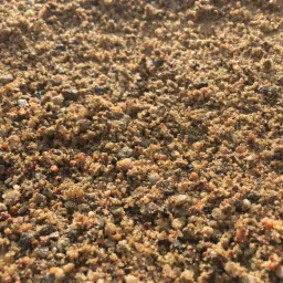 Bliskie ujęcie drobnego, mokrego materiału sypkiego o zróżnicowanej kolorystyce, od piaskowego po brązowy i czerwony, z widocznymi pojedynczymi kamyczkami i ziarnami, oświetlonego przez słońce.