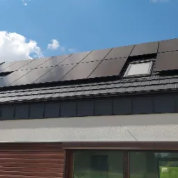 Panele fotowoltaiczne wkomponowane w dachówkę na nowoczesnym domu z oknami dachowymi, widoczne brązowe rolety okienne i błękitne niebo z chmurami.