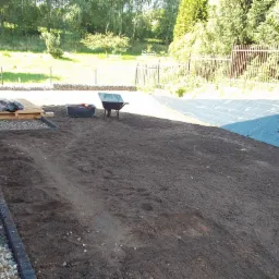 Przygotowany teren pod budowę, wyrównana ziemia z obrzeżami z kostki, w tle trawa, drzewa, siatka ogrodzeniowa, taczka, ubijarka i narzędzia.