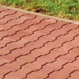 Czerwona nawierzchnia z kostki brukowej typu Behaton ułożona w zygzak, obrzeże z szarego betonu, trawa w tle.