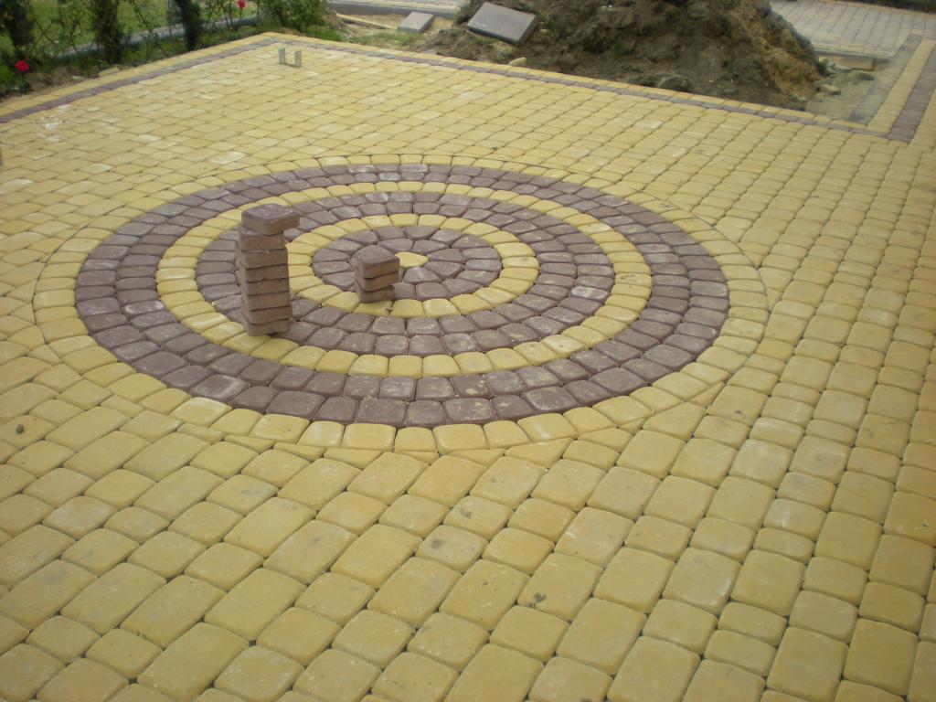 Ułożona kostka brukowa Behaton w dwóch kolorach, tworząca okrągły wzór na podjeździe, z ułożonymi pionowo kostkami w centrum, sugerującymi etap prac.