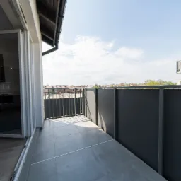 Nowoczesny balkon z szarą podłogą z płytek, częściowo otwartymi przesuwnymi drzwiami balkonowymi i widokiem na zabudowę miejską w tle, uchwycony w jasny, słoneczny dzień.