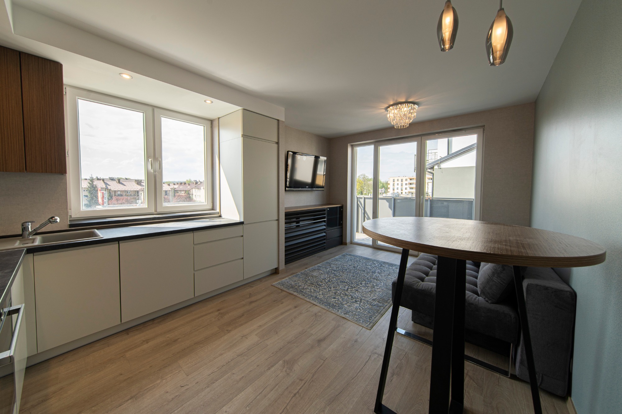 Jasne, nowoczesne wnętrze mieszkania z aneksem kuchennym, okrągłym stołem i balkonem, widok na zabudowę miejską przez duże okna.