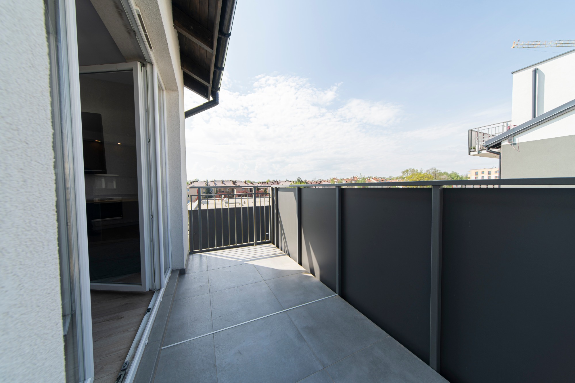 Nowoczesny balkon z szarą podłogą z płytek, częściowo otwartymi przesuwnymi drzwiami balkonowymi i widokiem na zabudowę miejską w tle, uchwycony w jasny, słoneczny dzień.
