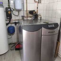 Szary kocioł Pellux z podłączoną instalacją hydrauliczną w kotłowni wyłożonej kafelkami, widoczny biały zbiornik wyrównawczy i niebieskie naczynie przeponowe.
