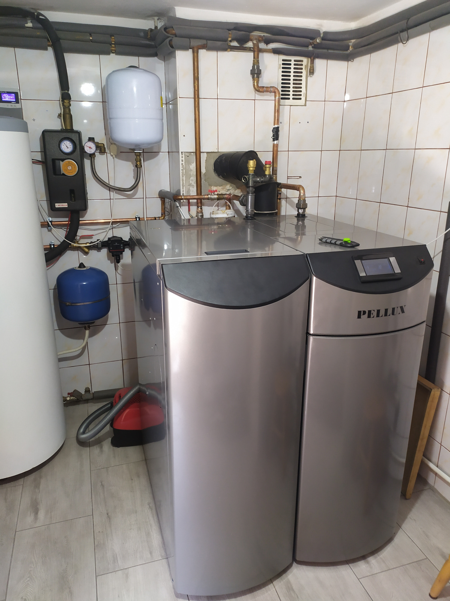 Szary kocioł Pellux z podłączoną instalacją hydrauliczną w kotłowni wyłożonej kafelkami, widoczny biały zbiornik wyrównawczy i niebieskie naczynie przeponowe.