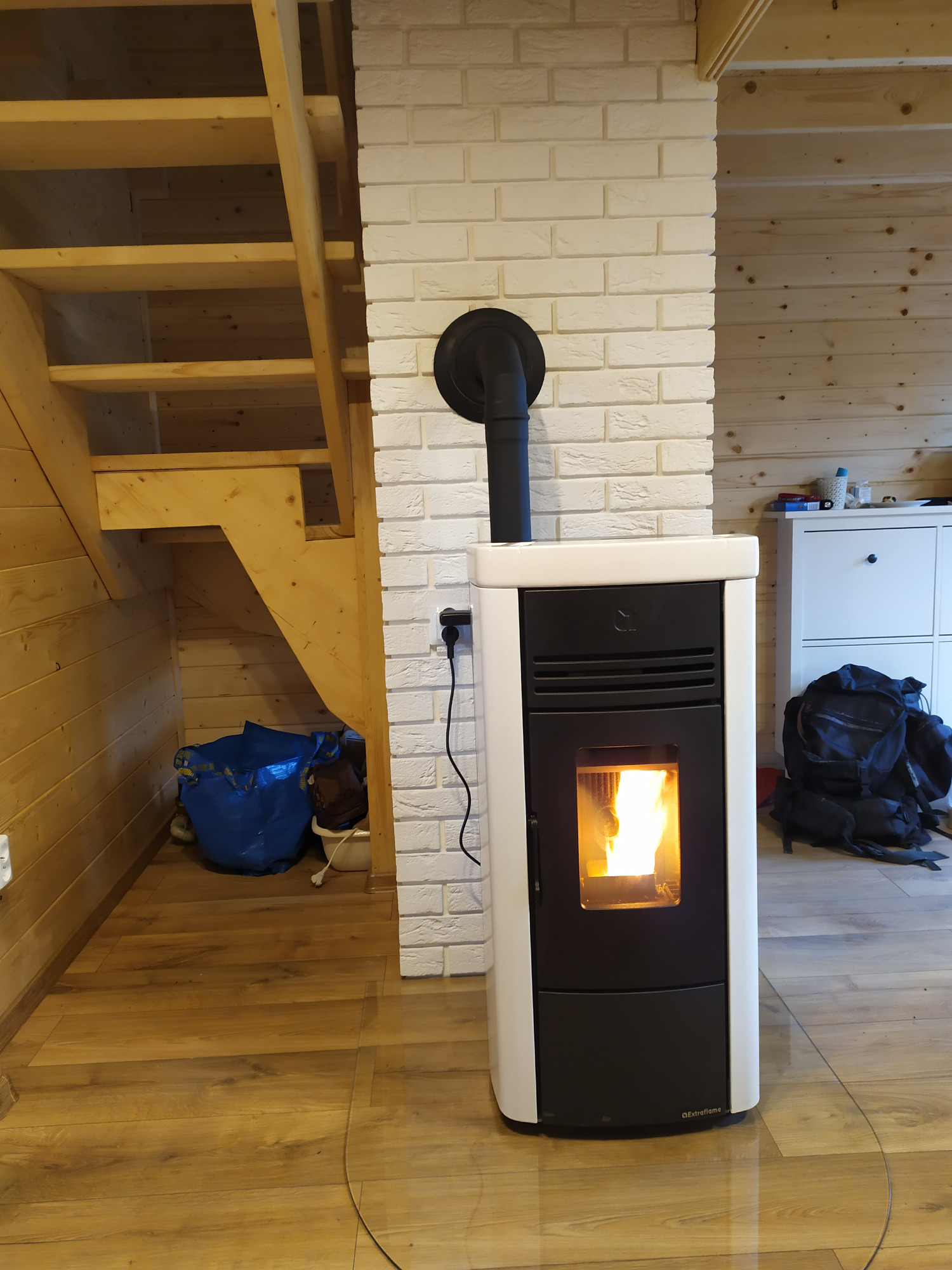 Nowoczesny piec na paliwo stałe, z płomieniem widocznym przez szybę, ustawiony w rogu pokoju z drewnianymi schodami i ścianą wyłożoną białymi cegłami.