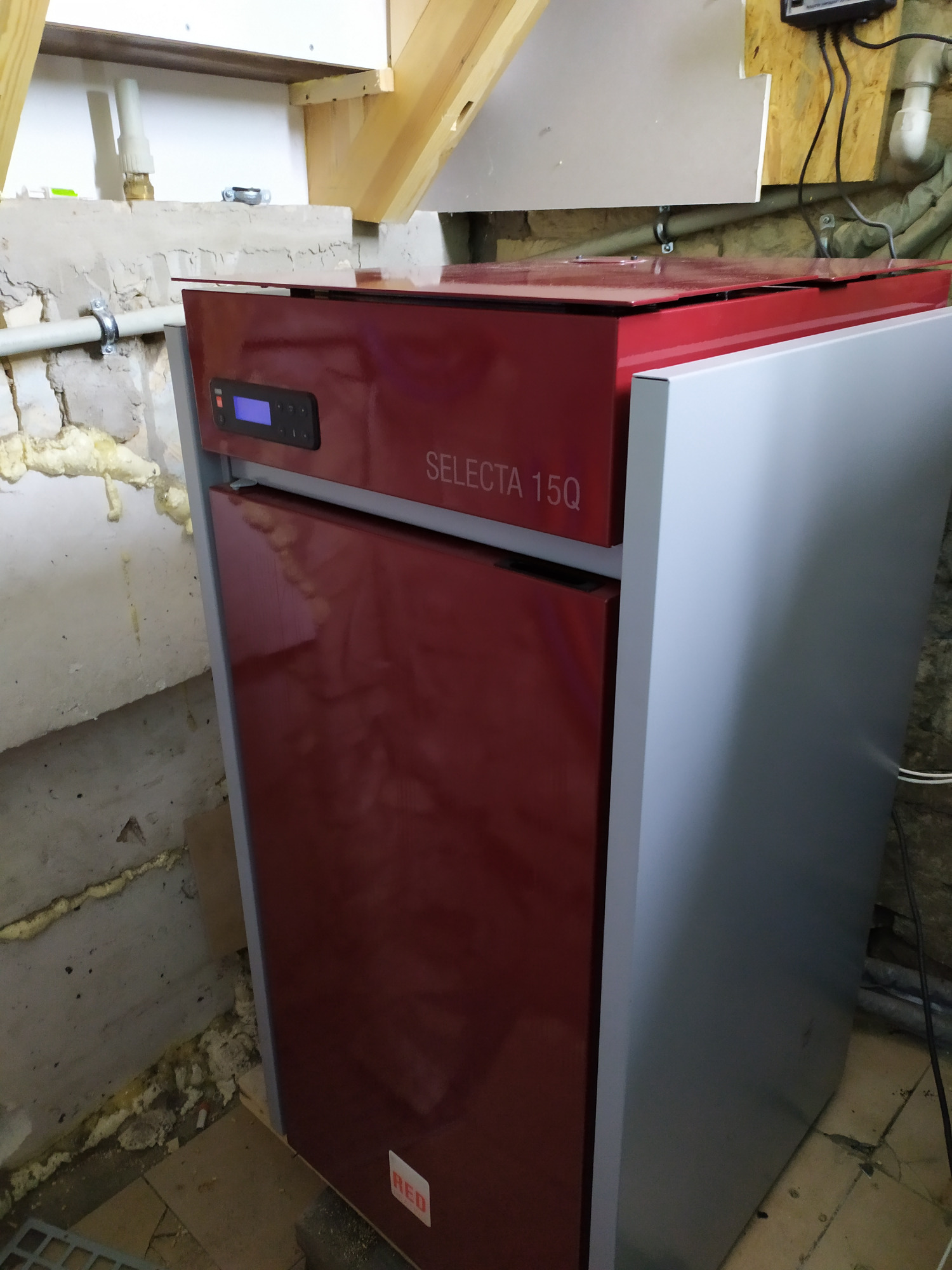 Czerwono-szary kocioł Selecta 15Q z wyświetlaczem, umieszczony w surowym wnętrzu z widoczną instalacją i ociepleniem piankowym na ścianie.