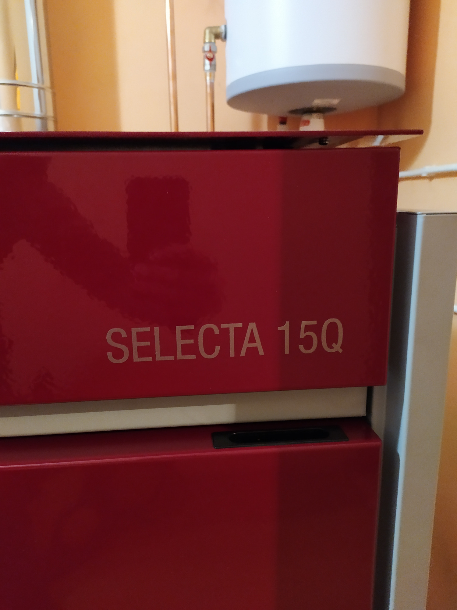 Czerwony piec SELECTA 15Q z widocznym zasobnikiem ciepłej wody i rurami miedzianymi w tle.