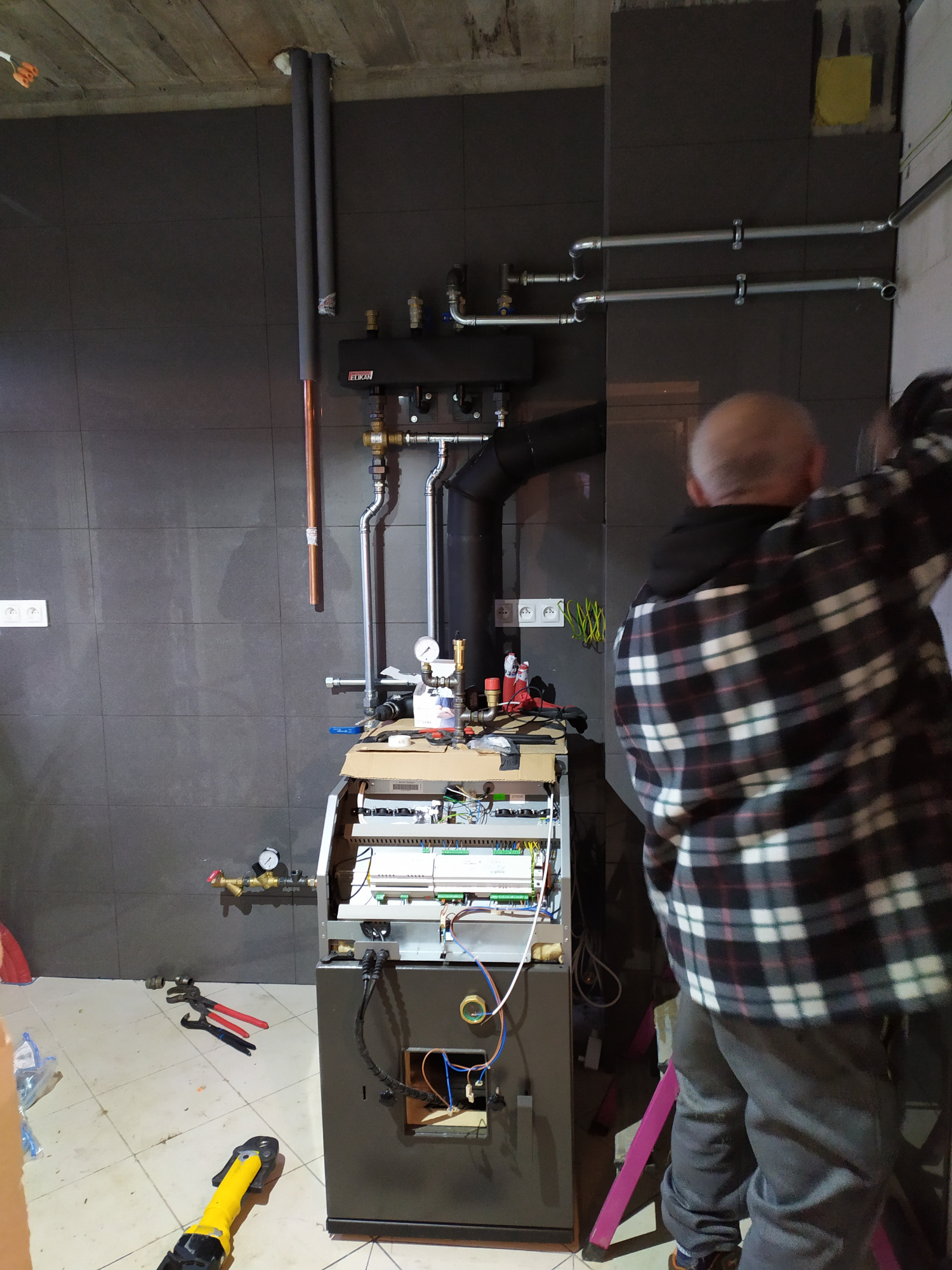 Instalacja kotła grzewczego z widocznymi rurami, manometrami i elementami sterującymi, instalator w koszuli w kratę w trakcie montażu na tle szarej ściany z płytek.