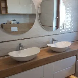 Nowoczesna łazienka z dwoma okrągłymi lustrami, umywalkami nablatowymi o organicznym kształcie, drewnianym blatem i białymi szafkami z oświetleniem pod szafkami, płytki ścienne w odcieniach...