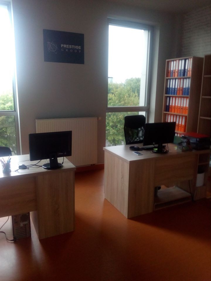Wnętrze biura z dwoma biurkami, komputerami, regałem z segregatorami i logo 'Prestige Group' na ścianie, widok z okna na zieleń.