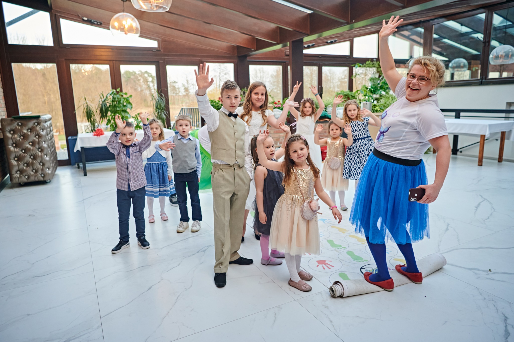 Animator w niebieskiej spódnicy i czerwonych balerinach, stojący na rozłożonej macie do gry dla dzieci, prowadzi grupę dzieci w różnym wieku, unoszących ręce w przestronnym, jasnym pomieszczeniu...