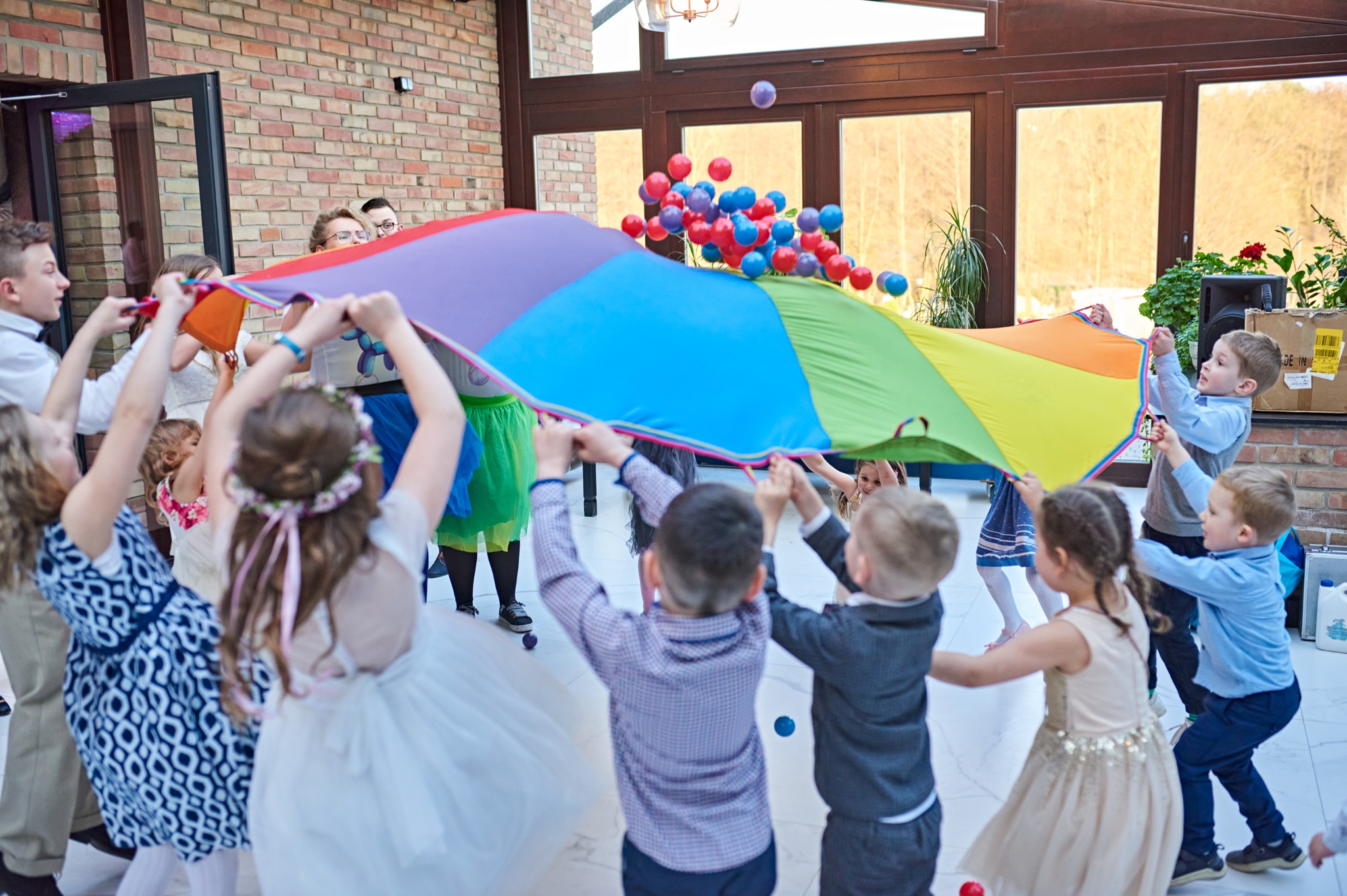 Grupa dzieci trzymająca kolorową chustę animacyjną, unosząc w górę czerwone i niebieskie piłeczki w jasnym pomieszczeniu z ceglanymi ścianami i dużymi oknami.