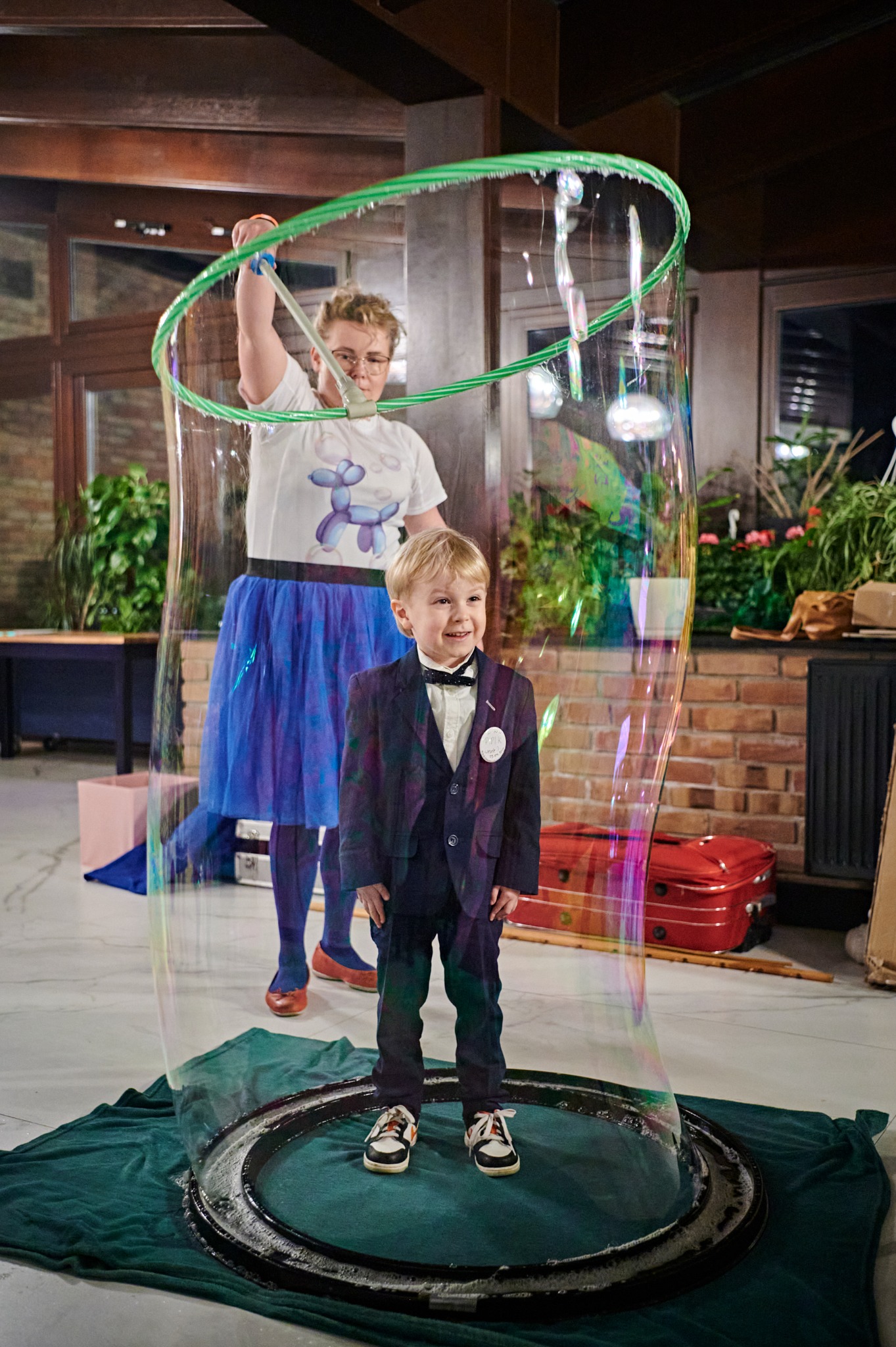 Uśmiechnięty chłopiec w garniturze stoi wewnątrz ogromnej bańki mydlanej, którą tworzy animator trzymający zielone obręcze; w tle rośliny doniczkowe i ceglana ściana.