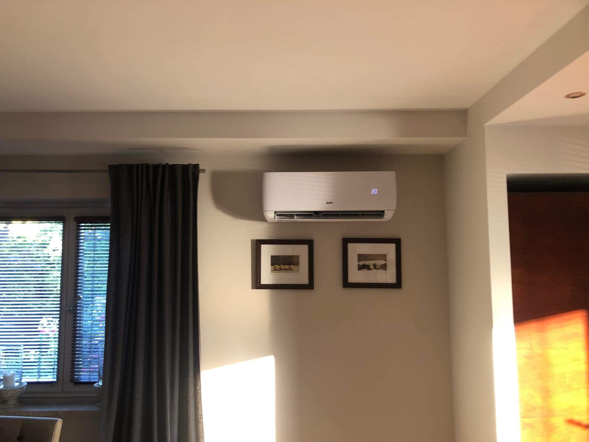Biały klimatyzator ścienny AUX z wyświetlaczem temperatury 20 stopni Celsjusza, zamontowany w salonie nad dwoma obrazkami w ciemnych ramkach.