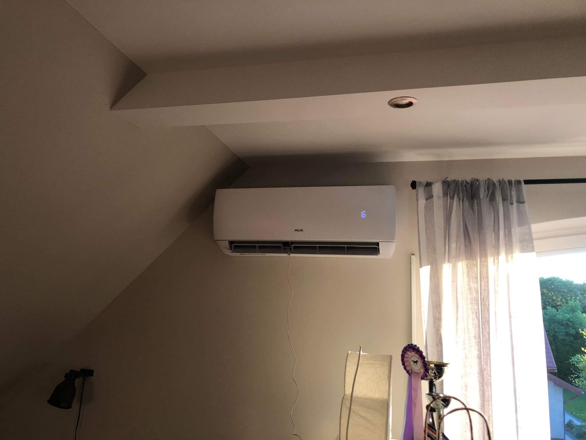 Biała jednostka klimatyzacyjna AUX zamontowana na ścianie w pokoju na poddaszu, widoczny kabel zasilający, obok okno z zasłonami i nagrody na stoliku.