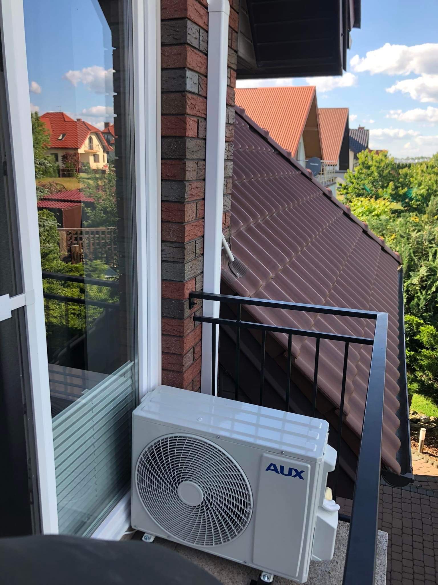 Zewnętrzna jednostka klimatyzacji AUX zamontowana na balkonie z widokiem na sąsiednie domy z czerwonymi dachami i zielenią, widoczne ceglane wykończenie balkonu i czarna balustrada.