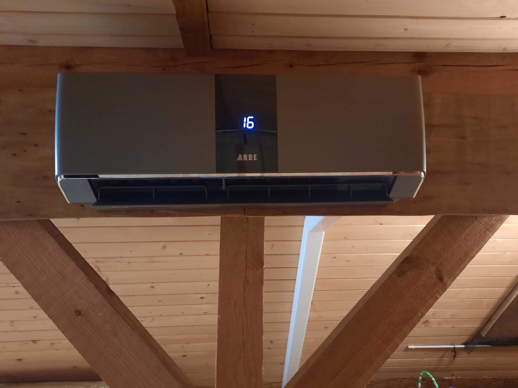 Szary klimatyzator z wyświetlaczem temperatury '16' zamontowany pod drewnianym stropem z widocznymi belkami.