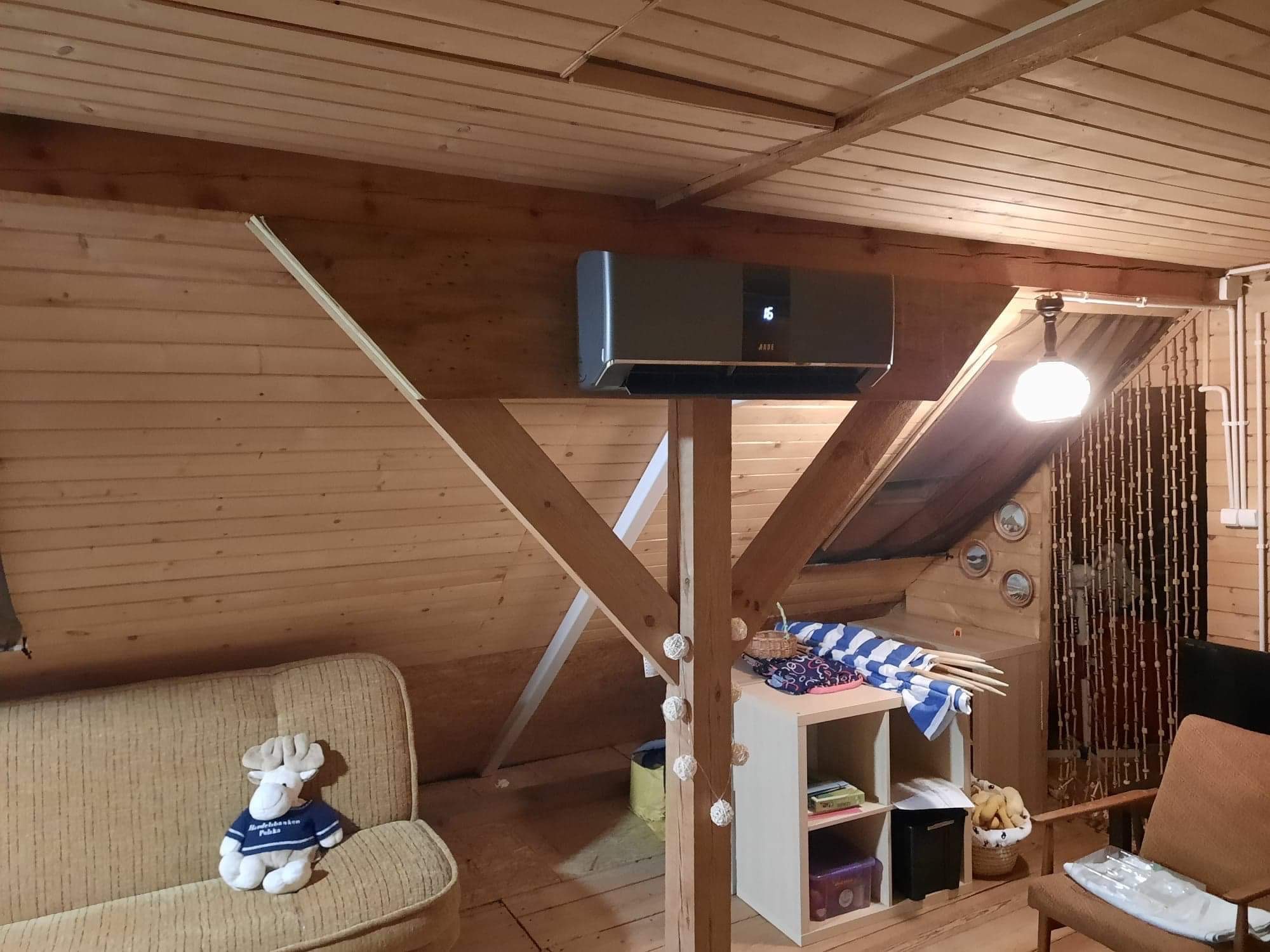 Szara klimatyzacja na drewnianej ścianie poddasza, wsparta konstrukcją z belek, widok na przytulne wnętrze z sofą i regałem.