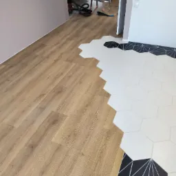 Połączenie paneli podłogowych z heksagonalnymi płytkami w geometryczny wzór. Widoczny fragment ściany i drabina w tle. Nietypowe zestawienie materiałów.