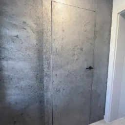 Ukryte drzwi z czarną klamką, wtopione w szarą ścianę o teksturze kamienia. Minimalistyczny design, nowoczesne wykończenie wnętrza, subtelne oświetlenie.