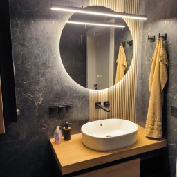 BestBud - Nowoczesna łazienka z okrągłym lustrem LED, umywalką nablatową na drewnianym blacie i czarnymi płytkami imitującymi marmur. Widoczne detale: dozownik mydła i ręcznik.
