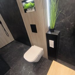 BestBud - Nowoczesna łazienka z wiszącą toaletą, czarnymi płytkami imitującymi marmur i dekoracyjnymi lamelami. Wnęka z roślinami i wysoka szafka z ręcznikiem dodają elegancji.