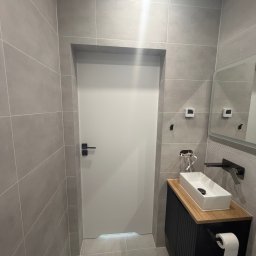 BestBud - Nowoczesna toaleta z szarymi, wielkoformatowymi płytkami na ścianach i podłodze. Białe drzwi z czarną klamką, umywalka nablatowa na drewnianym blacie i czarnej szafce. Widoczne lustro, termostat...