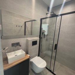 BestBud - Nowoczesna łazienka z szarymi płytkami, w tym teksturowanymi, białym umywalką na drewnianym blacie, czarną armaturą i kabiną prysznicową z czarnymi profilami. Minimalistyczny styl.
