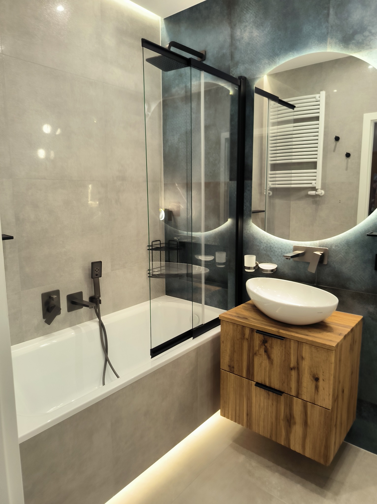 Nowoczesna łazienka z wanną, kabiną prysznicową i umywalką na drewnianej szafce. Lustro z podświetleniem LED. Użyto płytek w odcieniach szarości i błękitu. Armatura w kolorze czarnym.
