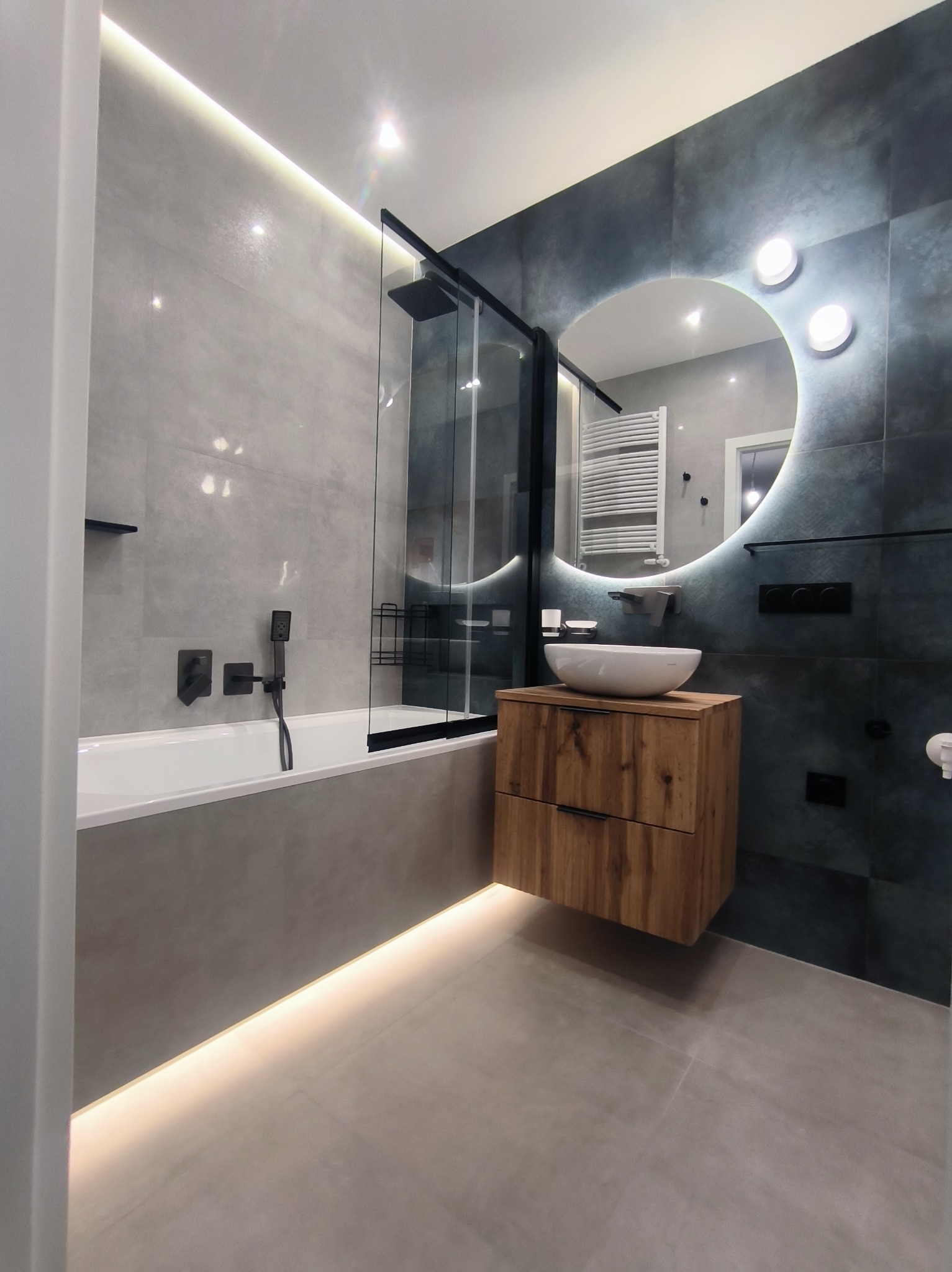 Nowoczesna łazienka z podświetlanym lustrem, drewnianą szafką pod umywalką i czarną armaturą. Minimalistyczny design z szarymi i granatowymi płytkami.