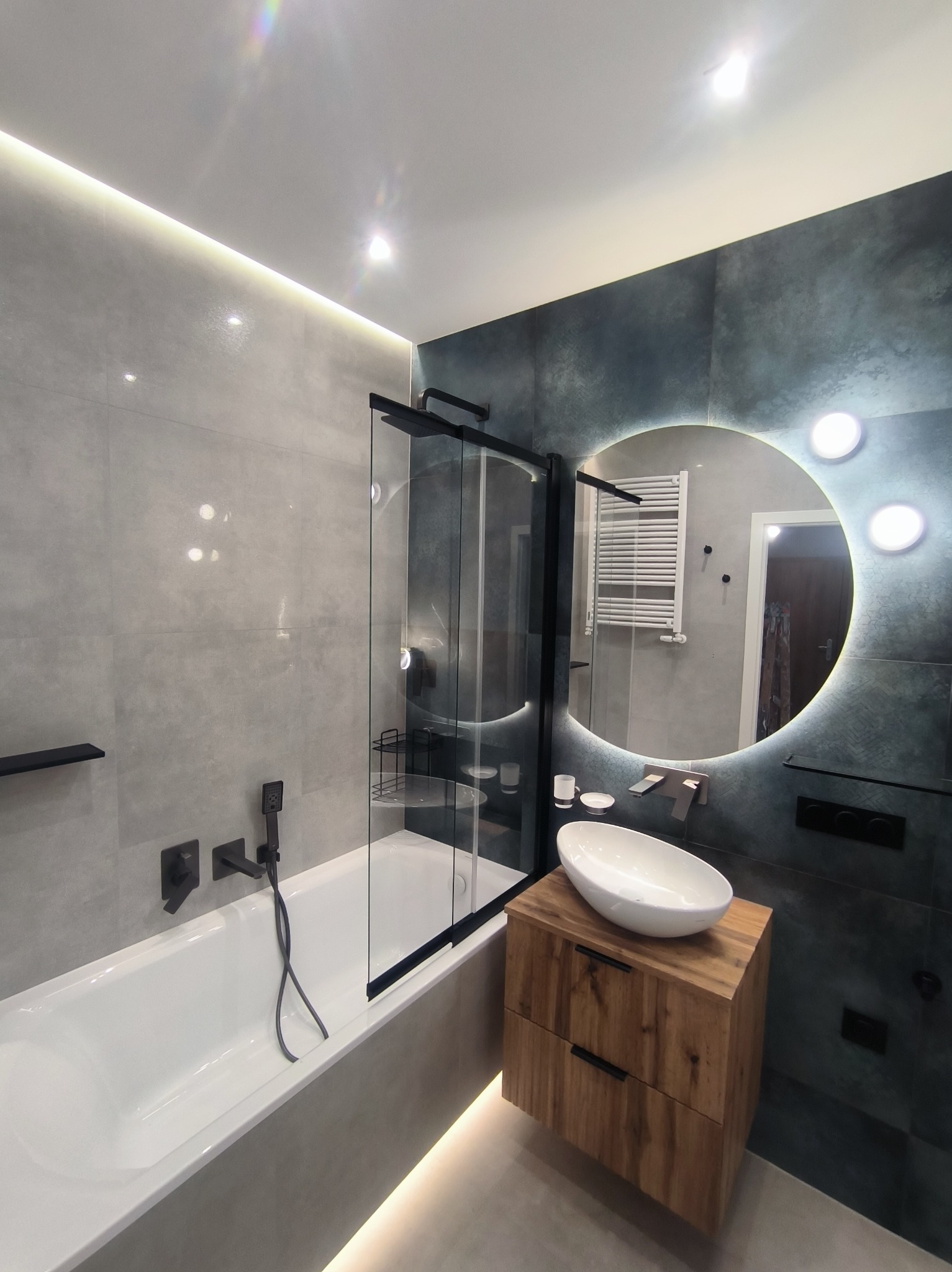 Nowoczesna łazienka z wanną i kabiną prysznicową oddzieloną szybą, umywalką na drewnianej szafce oraz okrągłym lustrem z podświetleniem LED.