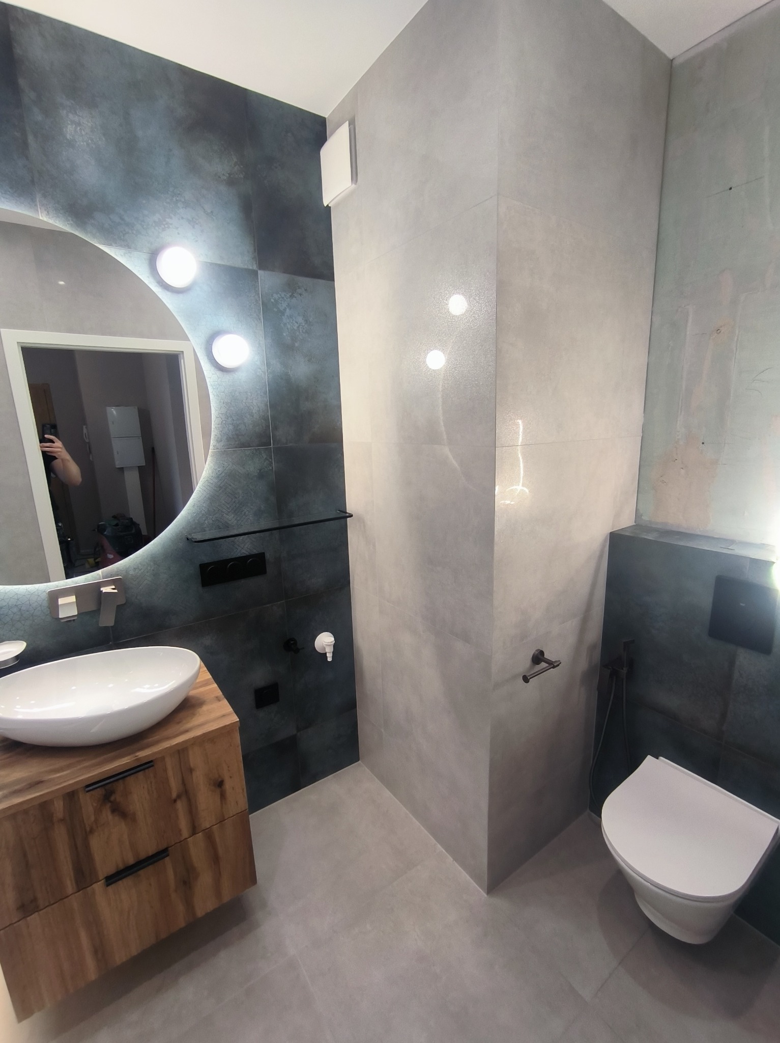 Nowoczesna łazienka z umywalką nablatową na drewnianej szafce, podświetlanym lustrem i szaro-grafitowymi płytkami. Widoczna toaleta podwieszana i minimalistyczne akcesoria.