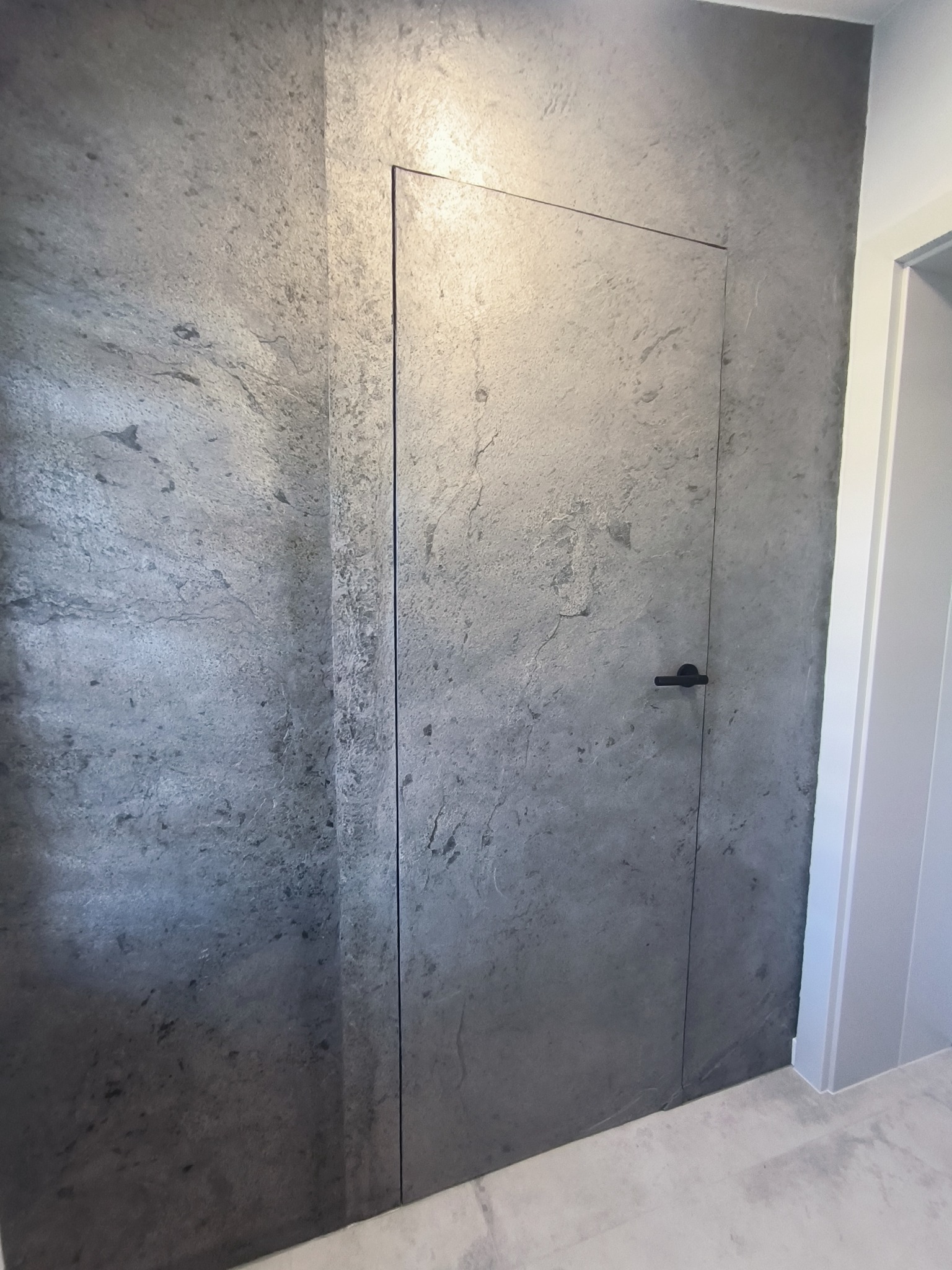 Ukryte drzwi z czarną klamką, wtopione w szarą ścianę o teksturze kamienia. Minimalistyczny design, nowoczesne wykończenie wnętrza, subtelne oświetlenie.