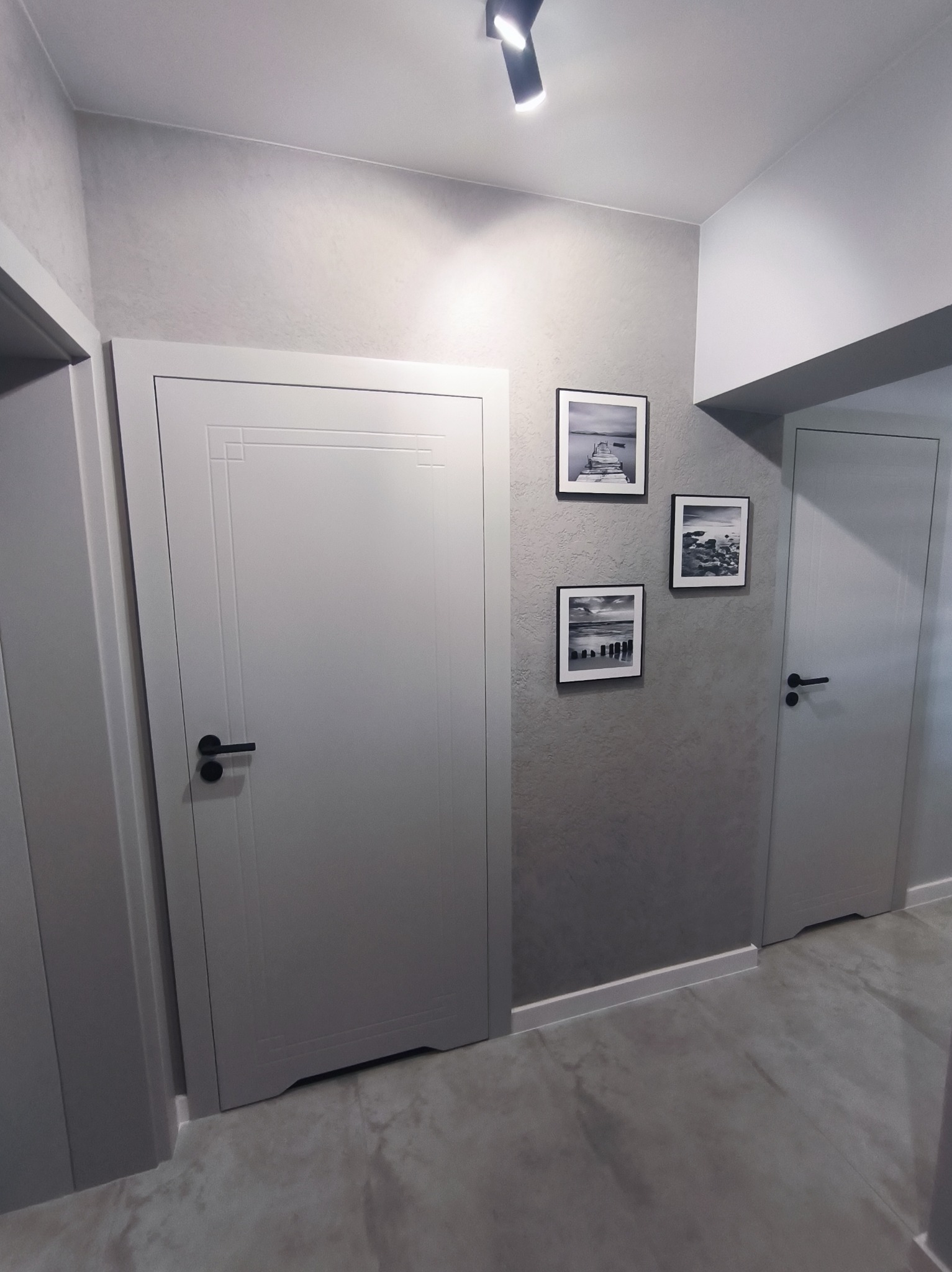 Szary korytarz po remoncie: dwie białe, zdobione drzwi z czarnymi klamkami, trzy czarno-białe zdjęcia na ścianie, szara podłoga i oświetlenie punktowe.