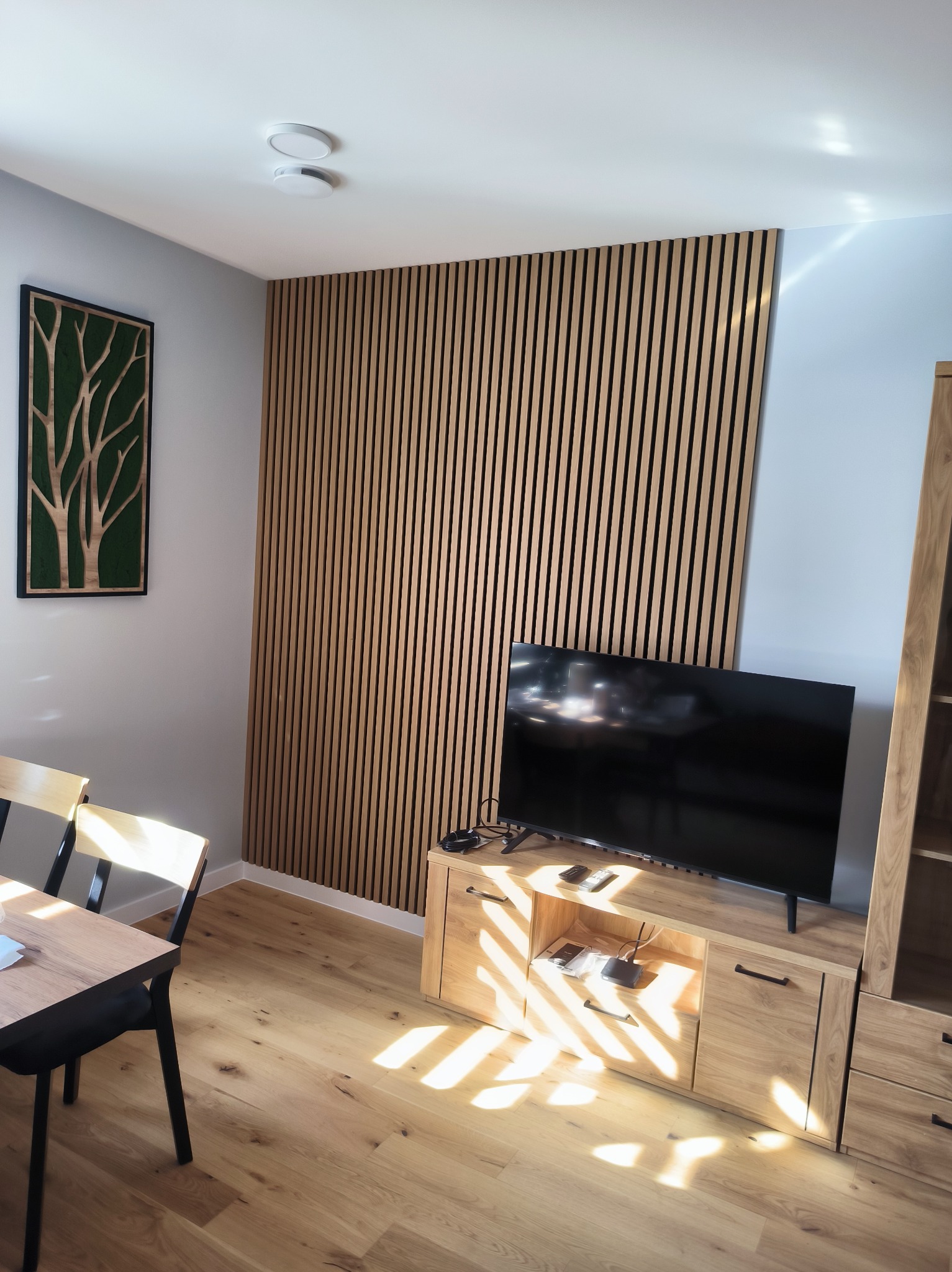 Nowoczesne wykończenie wnętrza z drewnianymi lamelami na ścianie, telewizorem na szafce RTV i drewnianą podłogą. Na ścianie wisi obraz z motywem drzewa.