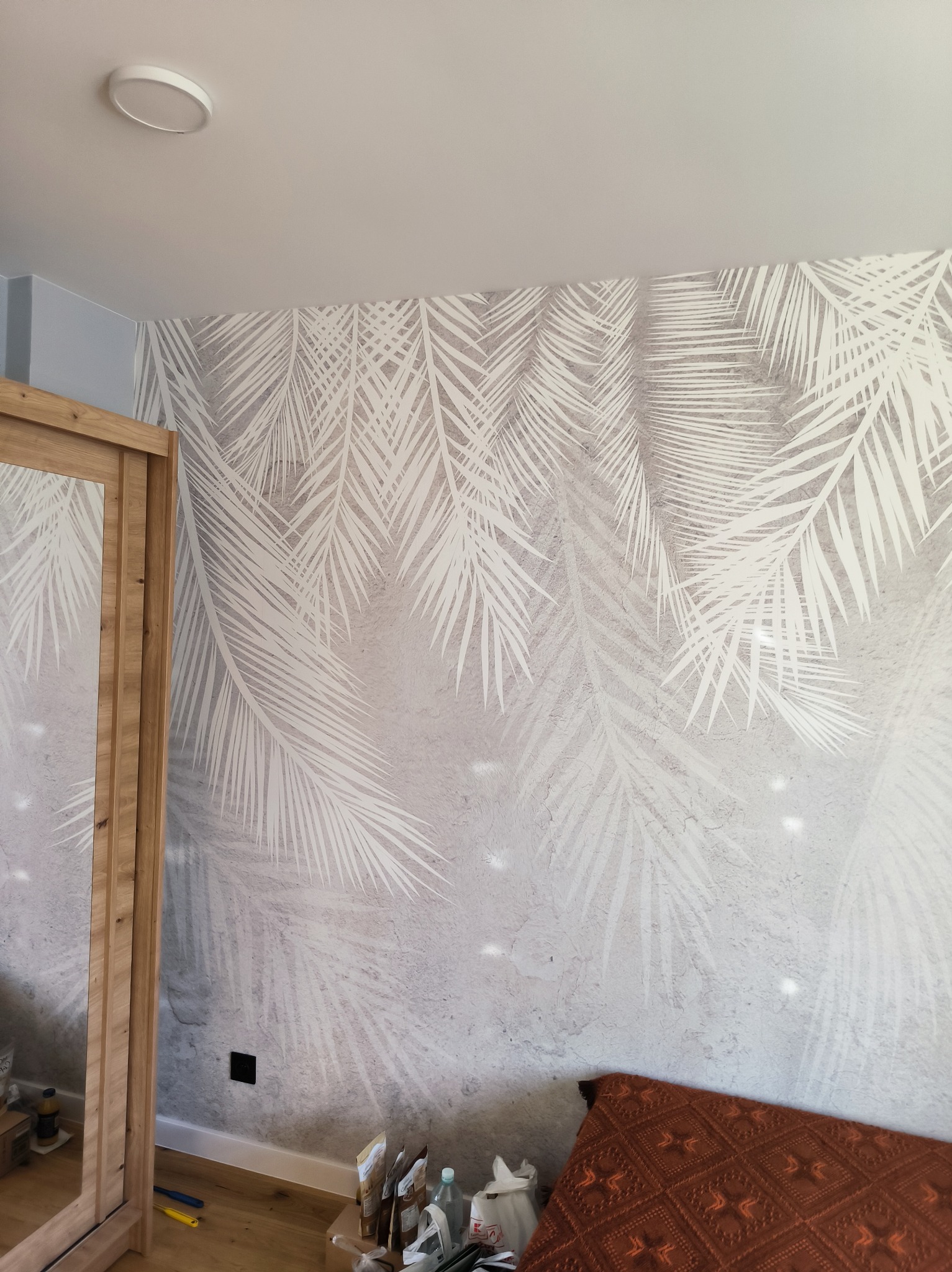Ściana z tapetą w białe liście palmowe na szarym tle, fragment drewnianej szafy z lustrem po lewej, widoczne gniazdko elektryczne i fragment podłogi.