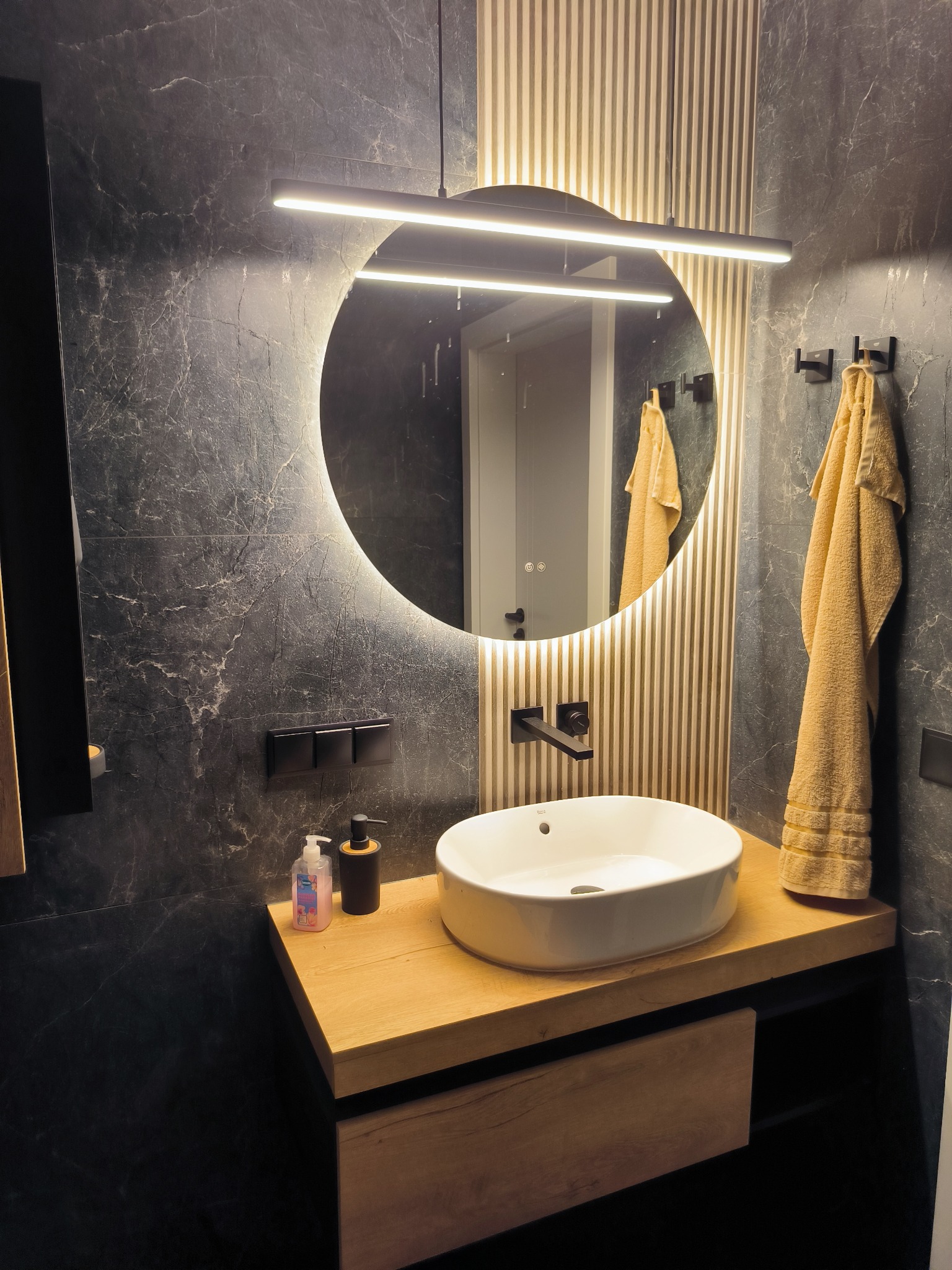 Nowoczesna łazienka z okrągłym lustrem LED, umywalką nablatową na drewnianym blacie i czarnymi płytkami imitującymi marmur. Widoczne detale: dozownik mydła i ręcznik.
