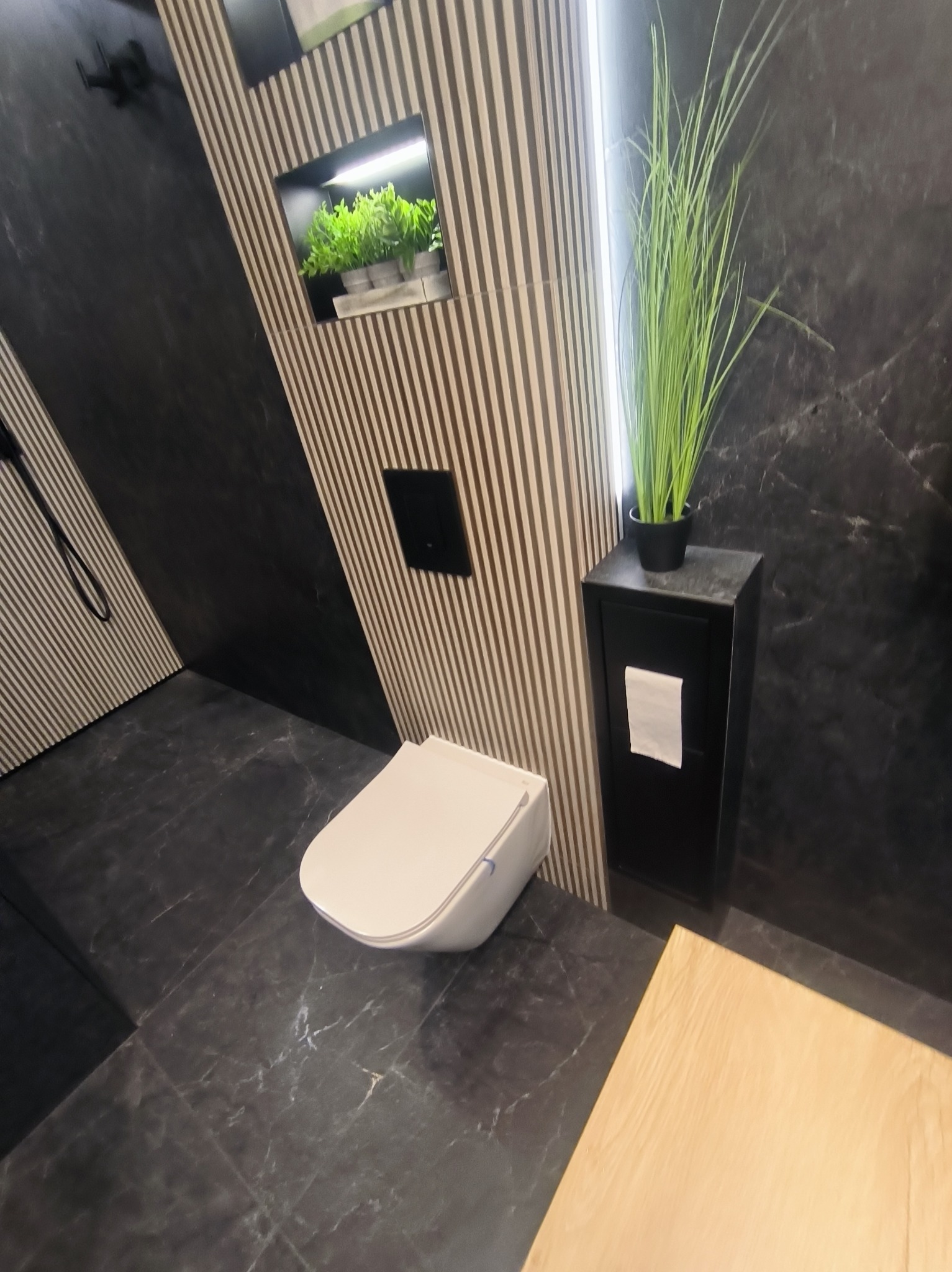 Nowoczesna łazienka z wiszącą toaletą, czarnymi płytkami imitującymi marmur i dekoracyjnymi lamelami. Wnęka z roślinami i wysoka szafka z ręcznikiem dodają elegancji.