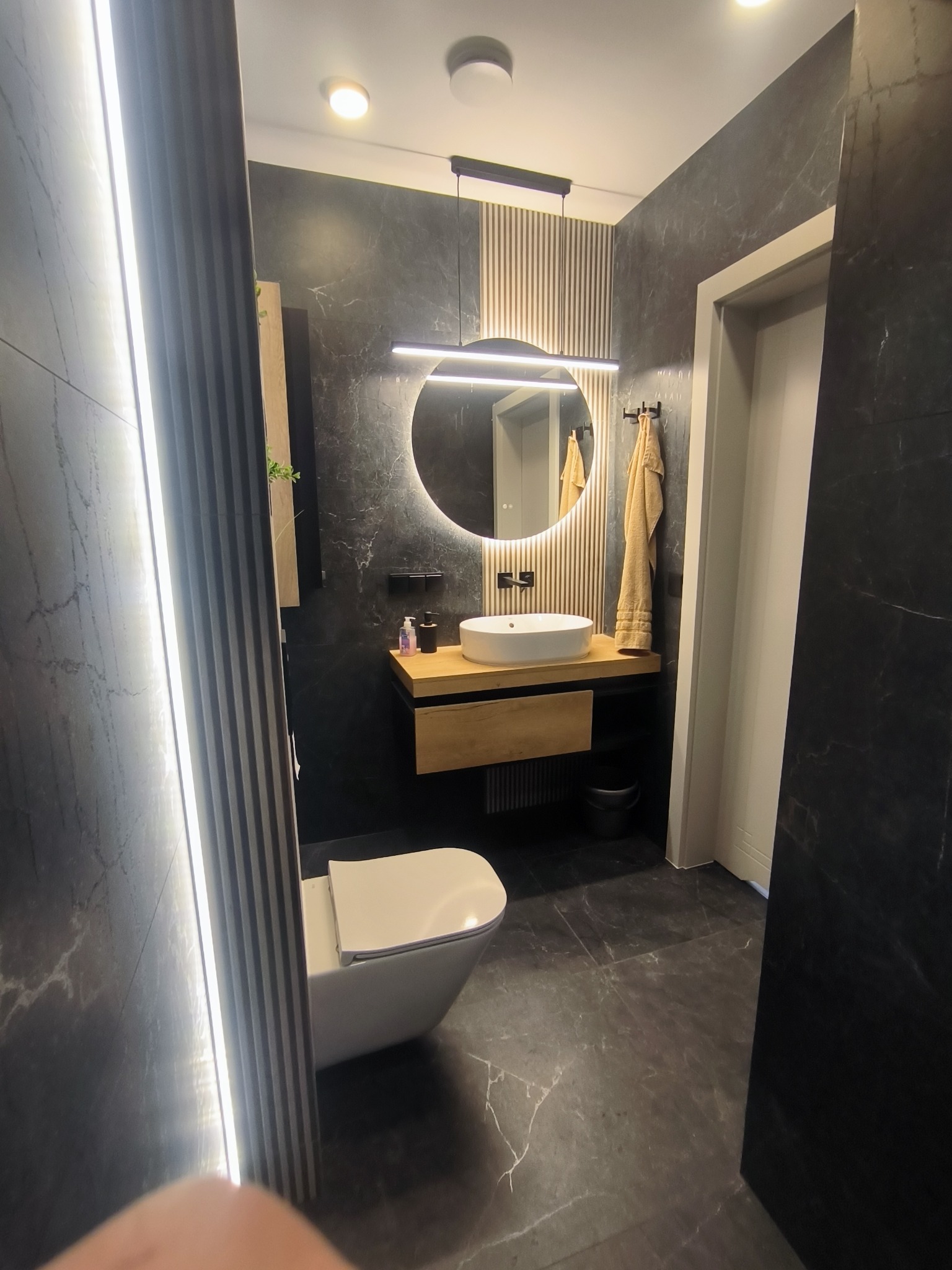 Nowoczesna łazienka z czarnymi płytkami imitującymi marmur, okrągłym lustrem z podświetleniem LED i drewnianą szafką pod umywalką. Minimalistyczny design.